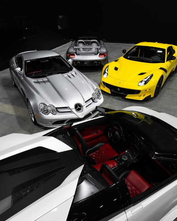 Cinematic, top-down view of a luxury automotive collection in a blacked-out showroom, including a silver Mercedes-Benz SLR McLaren, silver Porsche Carrera GT, and a bright yellow Ferrari F12tdf. In the foreground, a white Lamborghini Murcielago Roadster with its top off reveals a detailed red quilted leather interior.